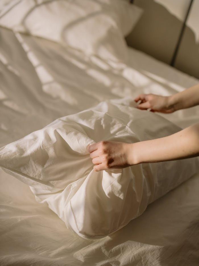 A close-up of hands adjusting a pillow on a neatly made bed, bathed in warm sunlight.